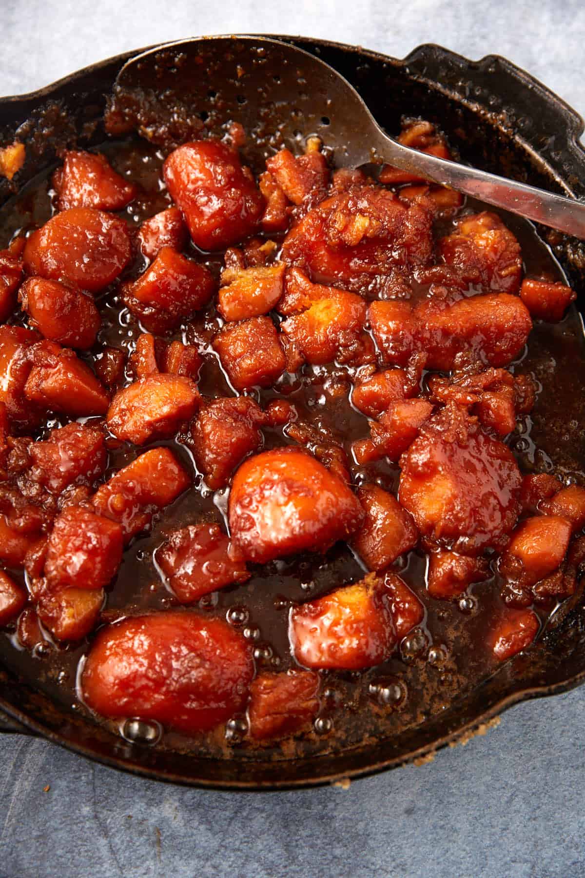 Chunks of caramelized pork belly coated in a glossy, rich brown sauce, served alongside sweet candied yams from a can in a black skillet with a metal spoon resting on the side.