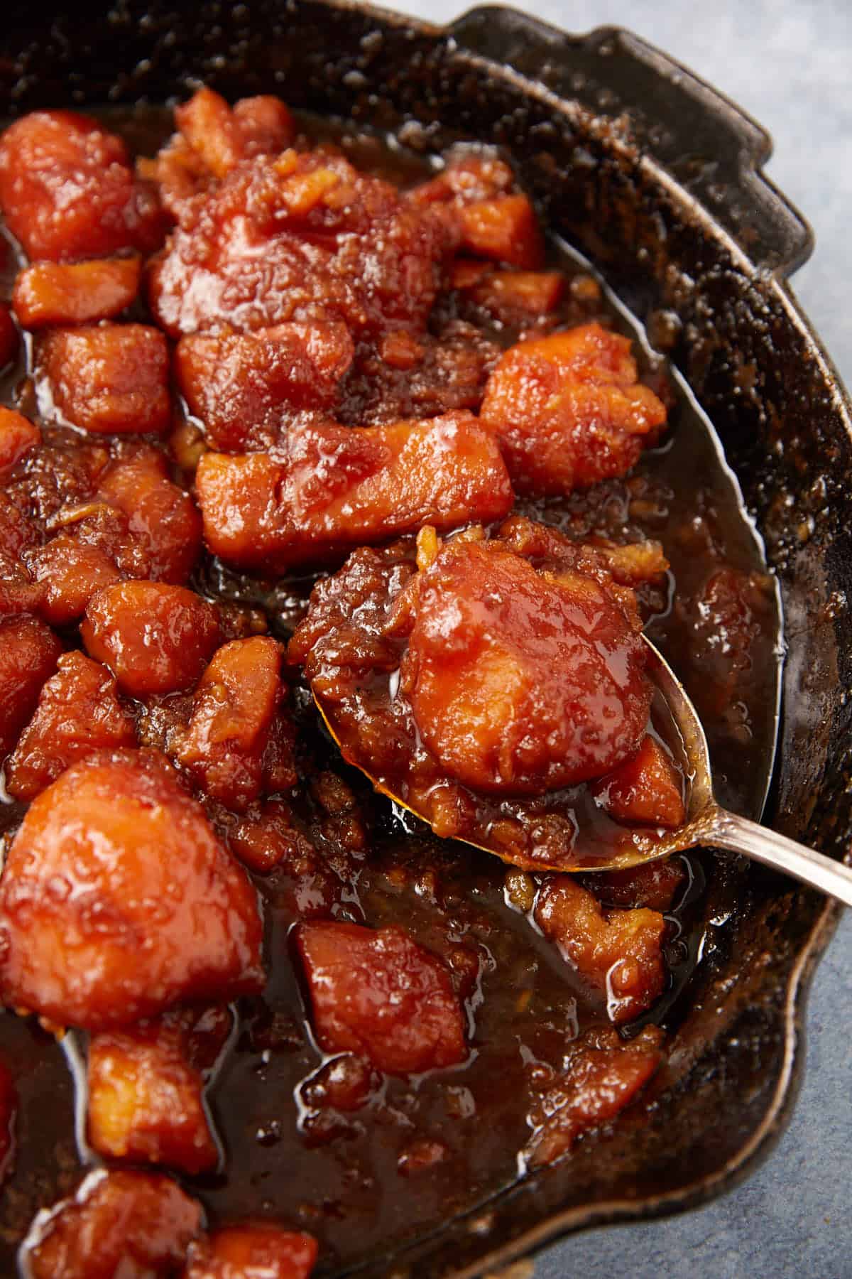 A close-up of baked candy yams in a skillet, coated in a glossy, caramelized brown sugar glaze, with a spoon lifting a portion.