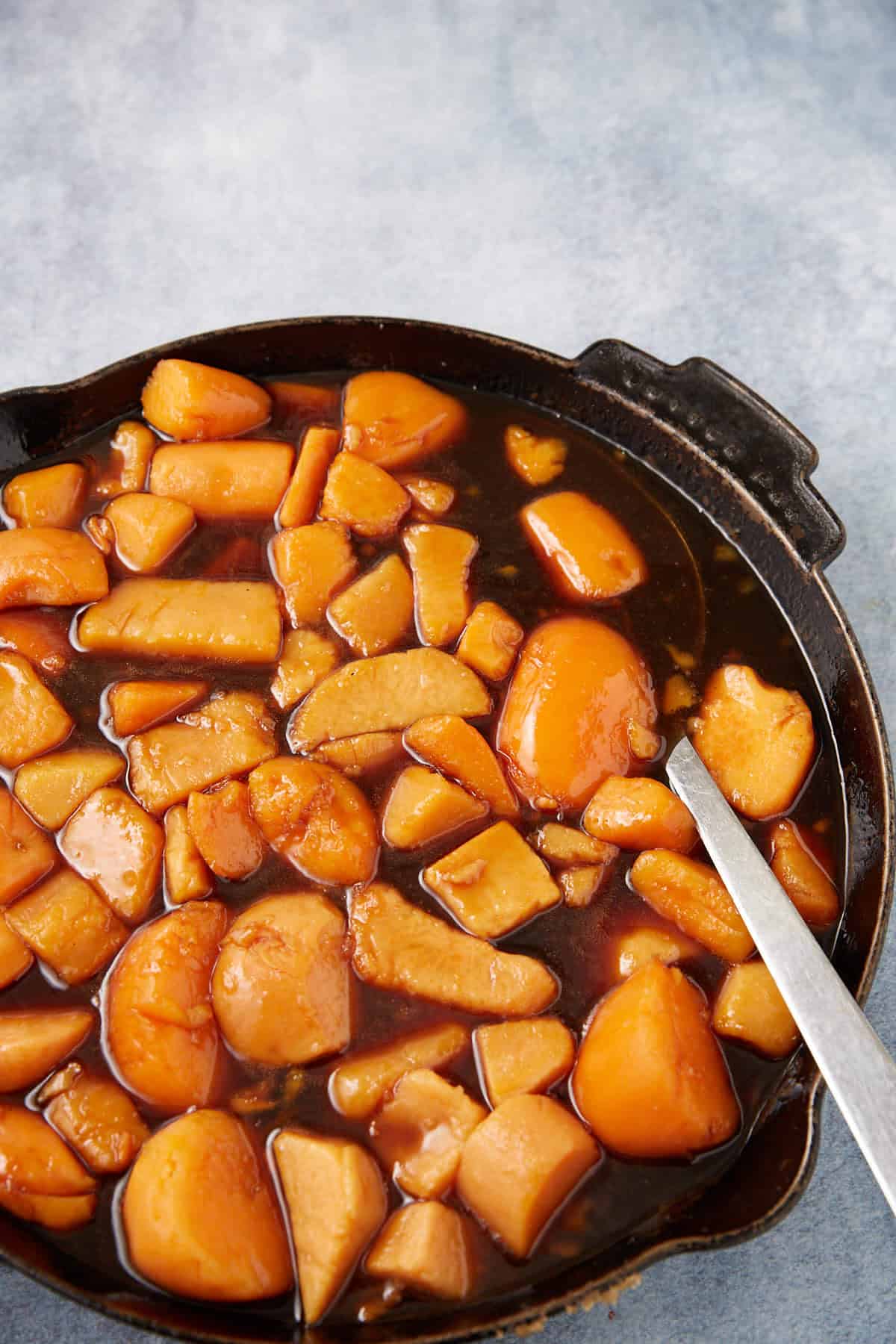 Slices of golden-brown cooked apples in a skillet, covered in a glossy syrupy sauce reminiscent of southern candied yams, with a metal spoon resting inside the pan on a light blue textured surface.