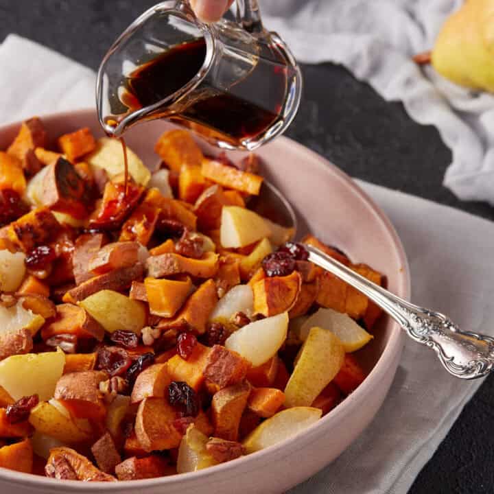 A serving bowl of roasted sweet potatoes with balsamic vinegar being poured over the top.