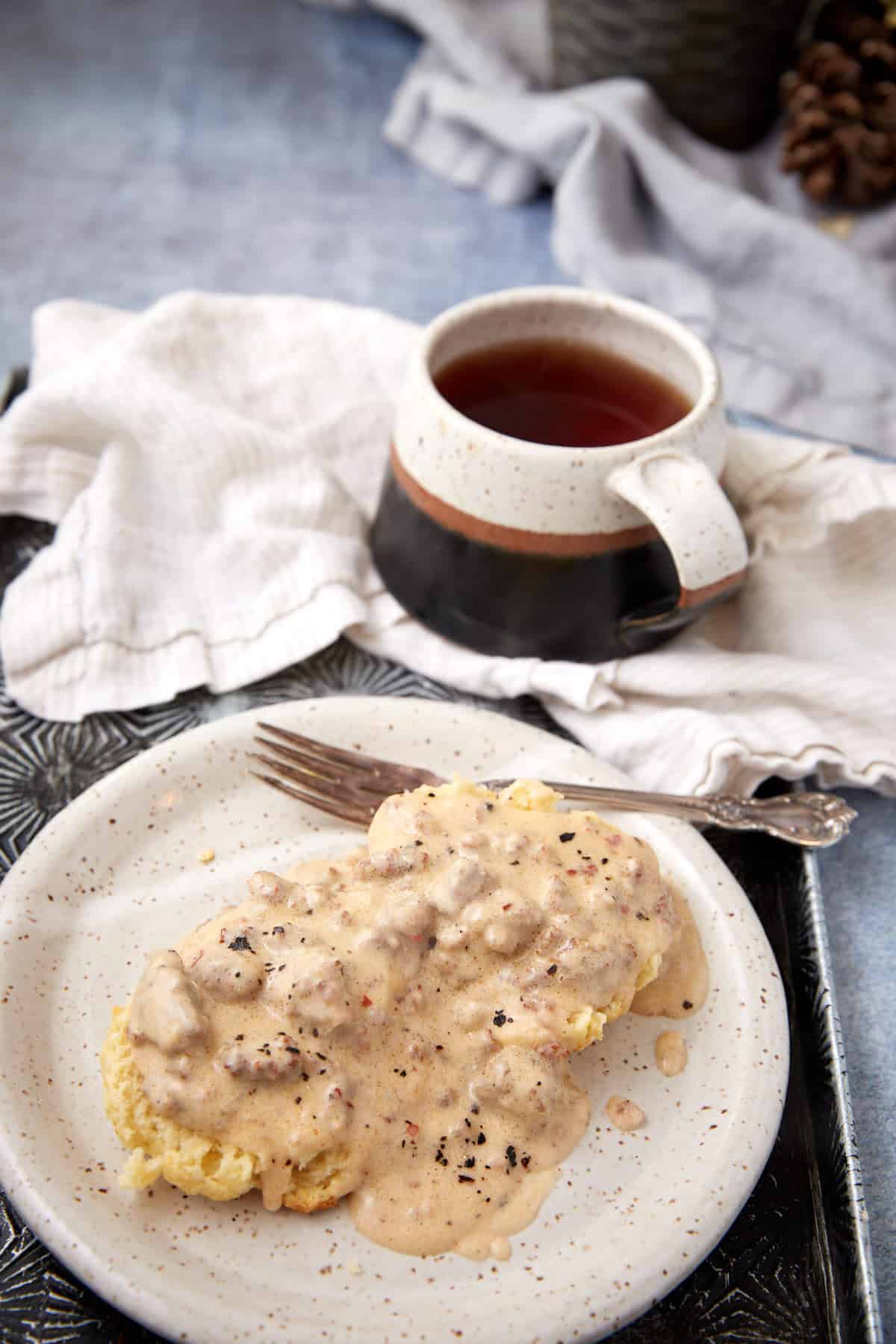 A plate with a biscuit smothered in savory sausage gravy sits beside a fork. In the background, a cup of tea or coffee rests on a textured tray alongside a folded cloth napkin.