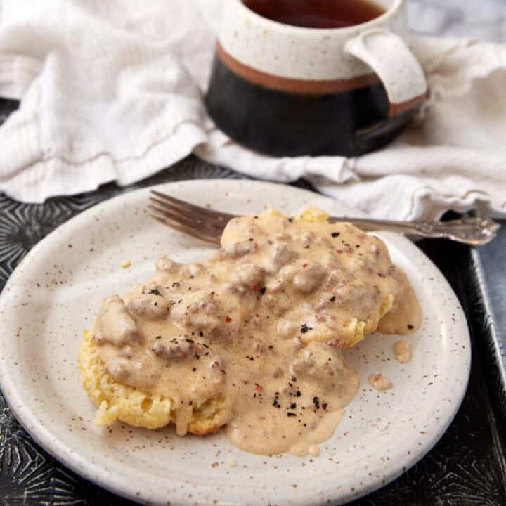 A white plate holds a biscuit topped with rich sausage gravy, with a fork beside it. In the background, there is a mug of dark tea and a folded white cloth napkin.