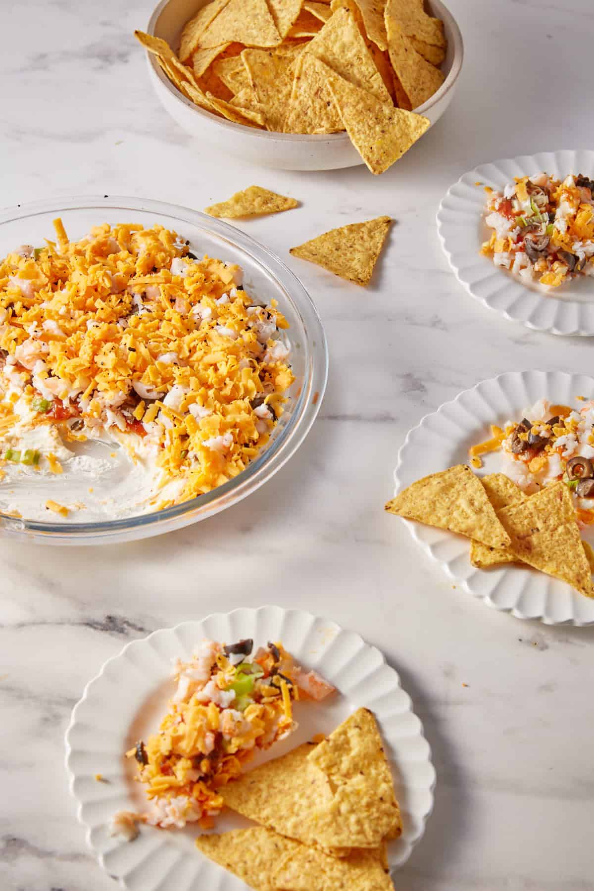 A glass dish with layered shrimp dip topped with shredded cheese is surrounded by tortilla chips. Some dip and chips are served on small white plates on a marble surface, with chips and crumbs scattered around.