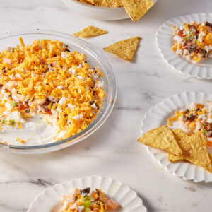 A glass dish with layered shrimp dip topped with shredded cheddar cheese sits on a marble surface. Three small plates nearby hold servings of the dip and tortilla chips. A bowl of chips is in the background.