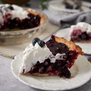 A slice of fresh blueberry pie topped with whipped cream and blueberries on a white plate, with the rest of the pie and another slice in the background—perfect inspiration for your next recipe for blueberry pie.