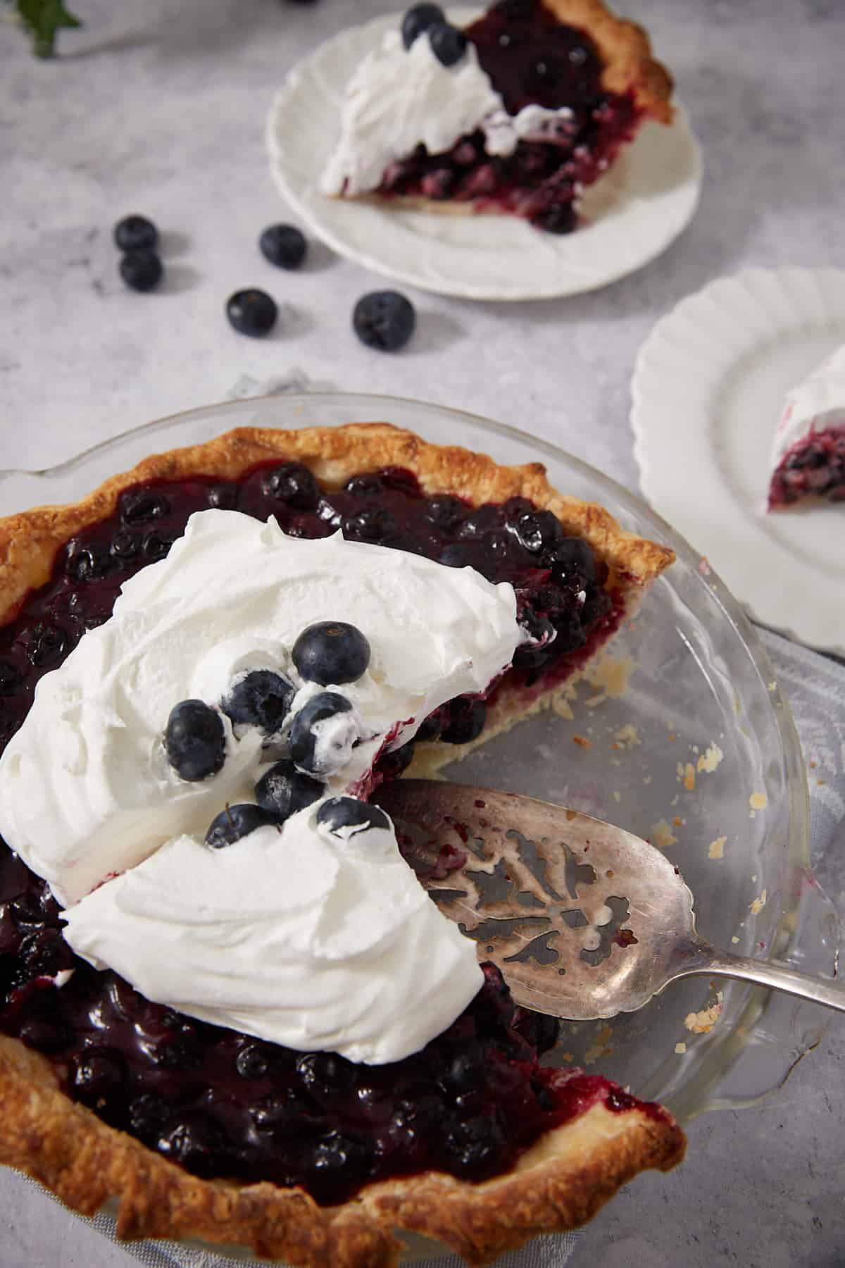 A fresh blueberry pie with a golden crust, topped with whipped cream and fresh blueberries. Try this easy recipe for fresh blueberry pie—a slice is served on a white plate in the background, and a pie server rests in the dish.