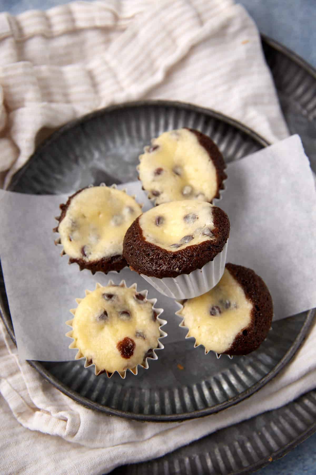 Four black bottom cupcakes with cream cheese and chocolate chip filling are arranged on a gray plate lined with parchment paper, set on a beige cloth napkin.