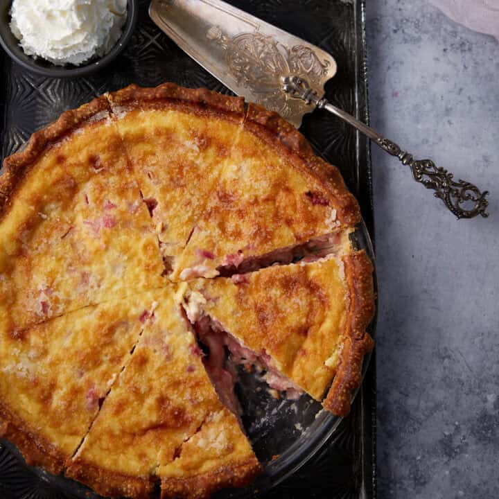 A rhubarb custard pie with a slice removed next to a bowl of whipped cream.