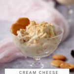 A glass dessert dish filled with creamy banana pudding with cream cheese is garnished with crumbled cookies and a whole cookie on the side. The background is soft and slightly blurred, and the text on the image reads "Cream Cheese Banana Pudding" and "butterandbaggage.com.
