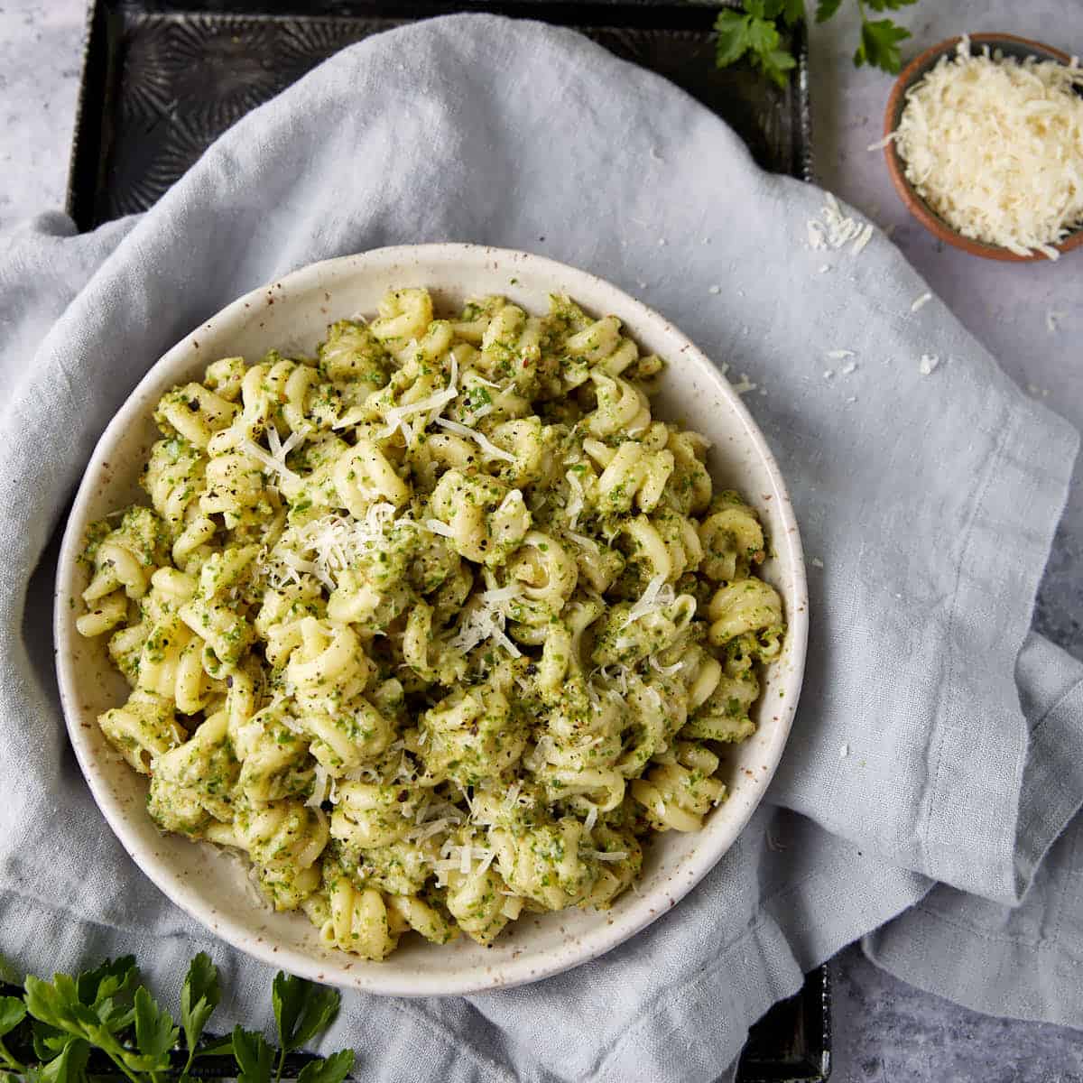 A bowl of pesto pasta with parsley next to parmesan cheese.