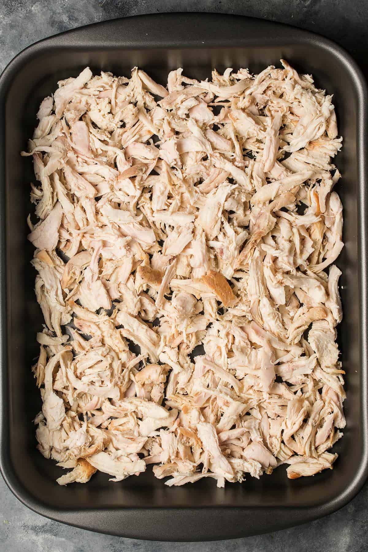 A baking pan filled with shredded cooked chicken, perfect for starting a classic Chicken Divan, spread evenly across the bottom on a dark countertop.
