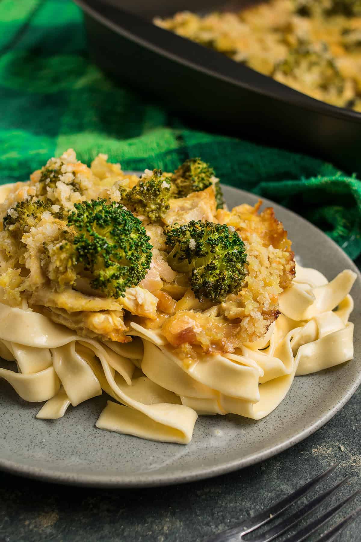 A plate of wide, flat pasta topped with a cheesy Chicken Divan broccoli and breadcrumb casserole, with a baking dish of the casserole in the background.