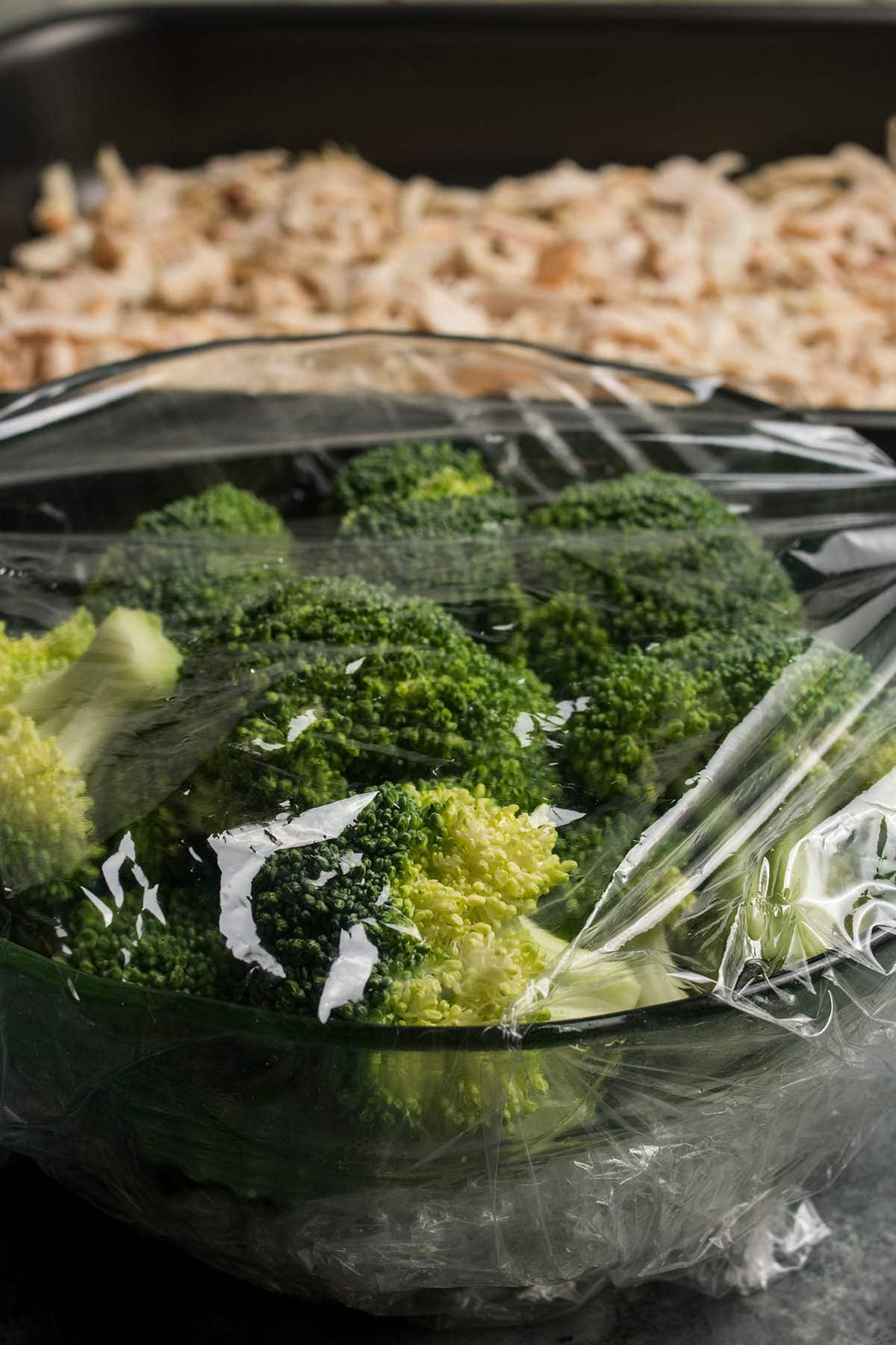 A bowl of fresh broccoli covered with plastic wrap is in the foreground, while a baking tray with shredded cooked chicken, perfect for Chicken Divan, is visible in the blurry background.