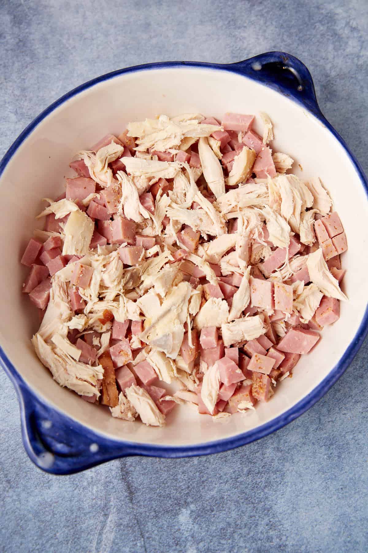 A white and blue bowl filled with shredded cooked chicken and diced ham—perfect for making a hearty chicken cordon bleu casserole—rests on a blue textured surface.