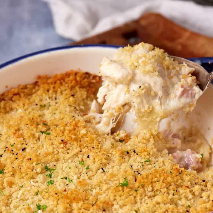 A spoon scooping out a creamy, cheesy chicken cordon bleu casserole topped with a golden, crispy breadcrumb crust from a baking dish.