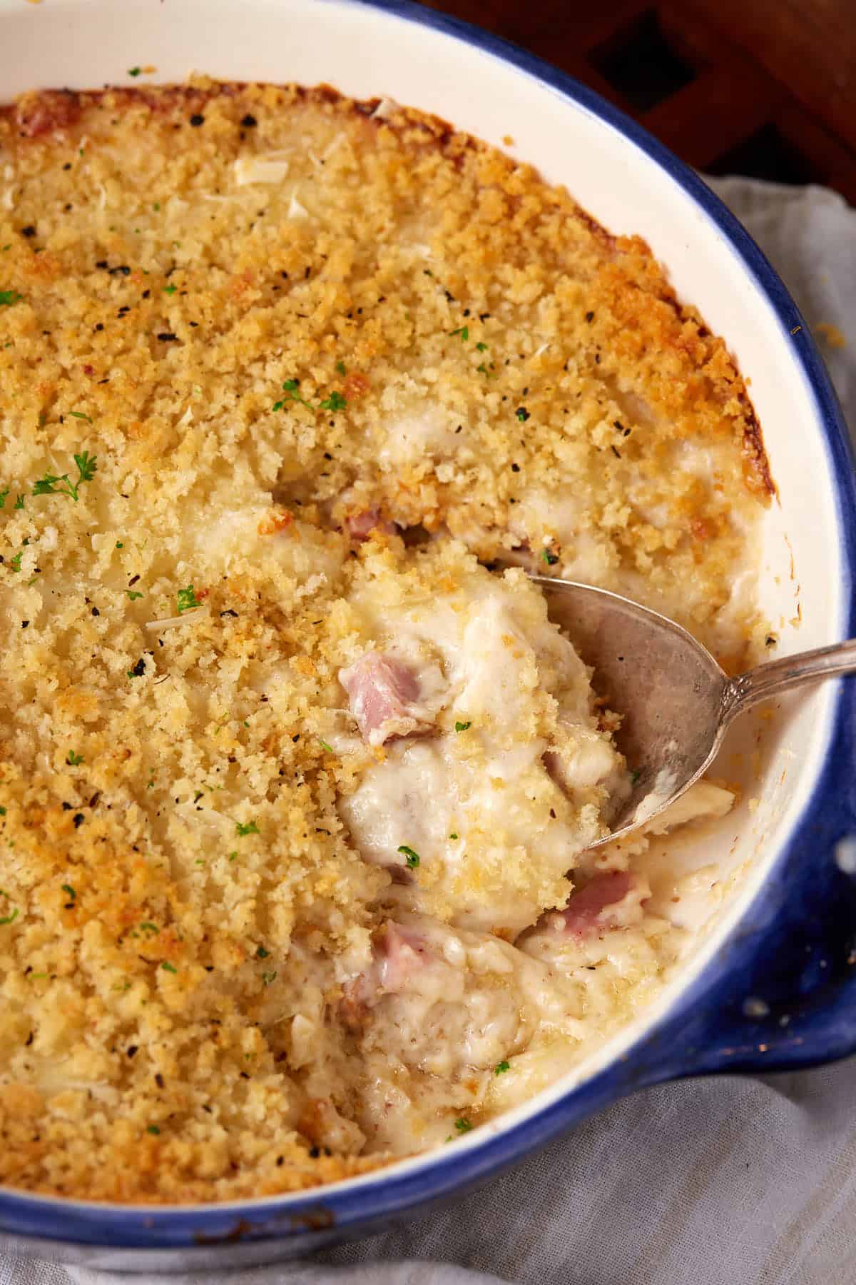 A close-up of a chicken cordon bleu casserole with a golden breadcrumb topping, showing a creamy, cheesy filling with chunks of ham being scooped out with a large spoon in a blue-rimmed baking dish.