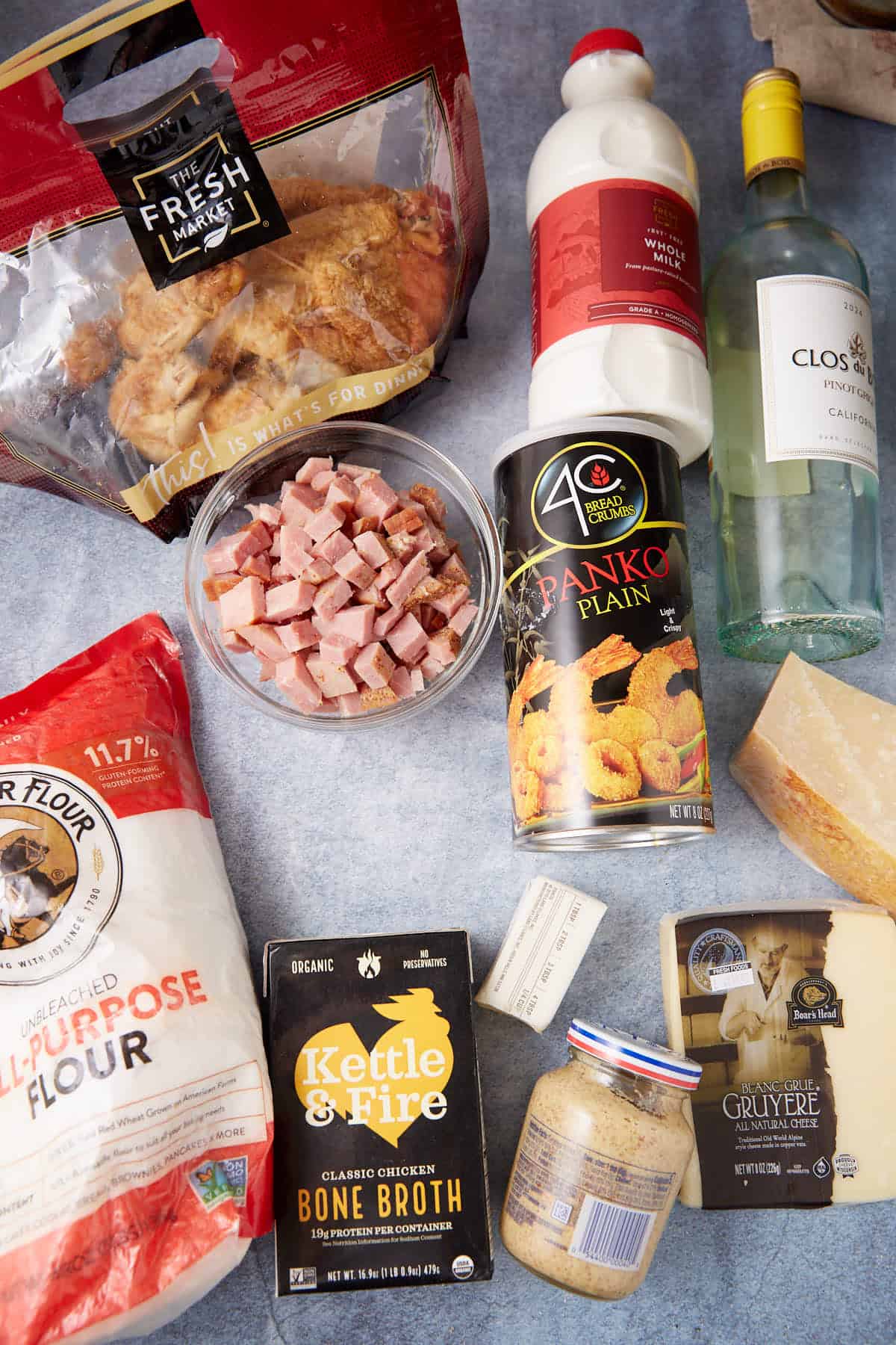 A variety of ingredients on a countertop, perfect for a chicken cordon bleu casserole—rotisserie chicken, diced ham, all-purpose flour, bone broth, plain panko, Parmesan and Gruyère cheeses, Dijon mustard, milk, and a bottle of white wine.