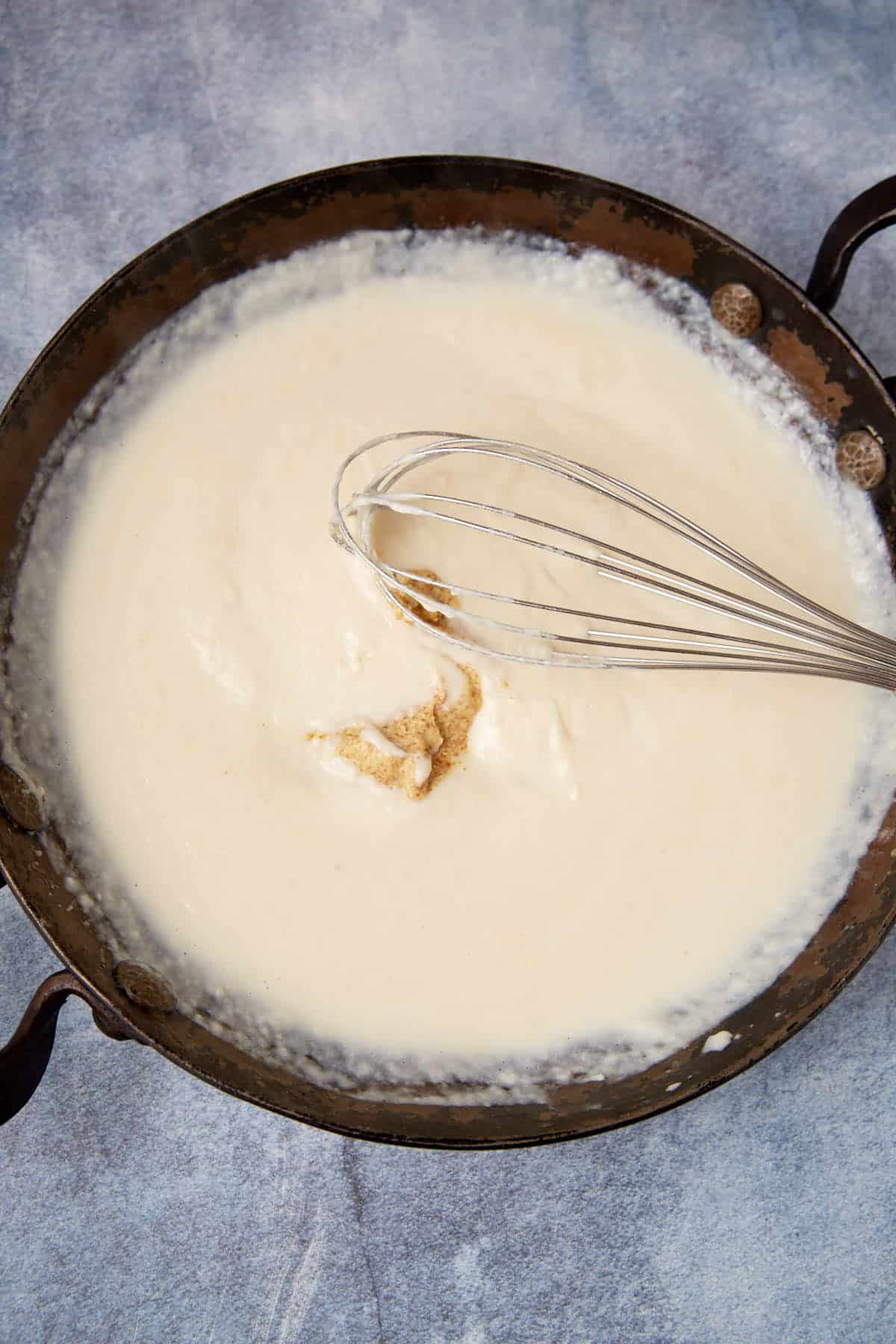 A metal whisk mixing a creamy white sauce for chicken cordon bleu casserole in a rustic, round pan on a blue-gray surface.