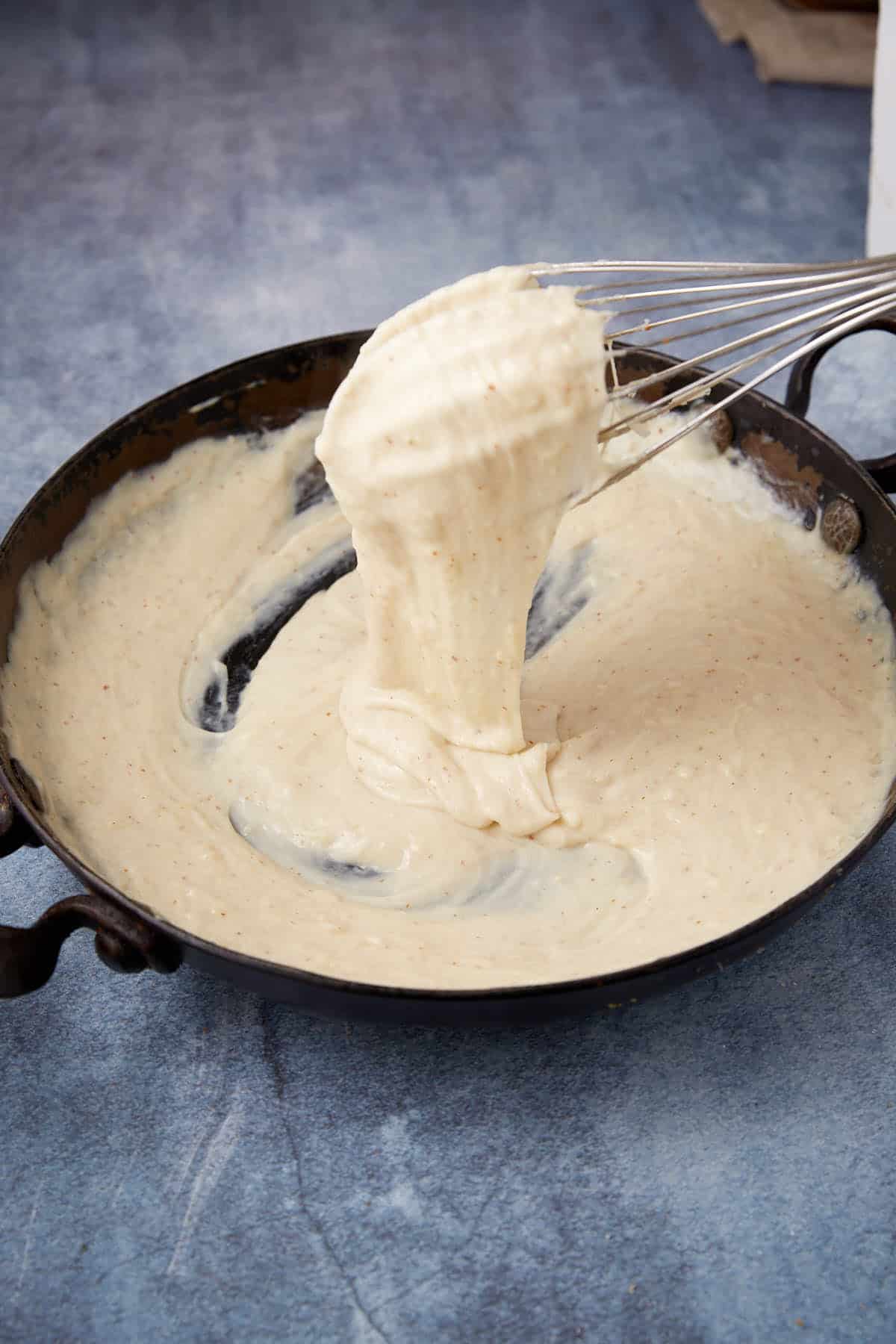 A whisk lifts thick, creamy béchamel sauce from a black skillet, perfect for layering in a chicken cordon bleu casserole. The sauce stretches between the whisk and pan against a textured blue background.