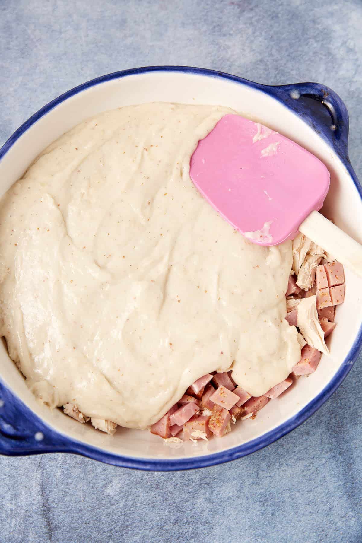 A blue casserole dish filled with chopped cooked meat and shredded chicken, partially covered with a thick layer of creamy white sauce being spread with a pink spatula—perfect for a hearty chicken cordon bleu casserole.