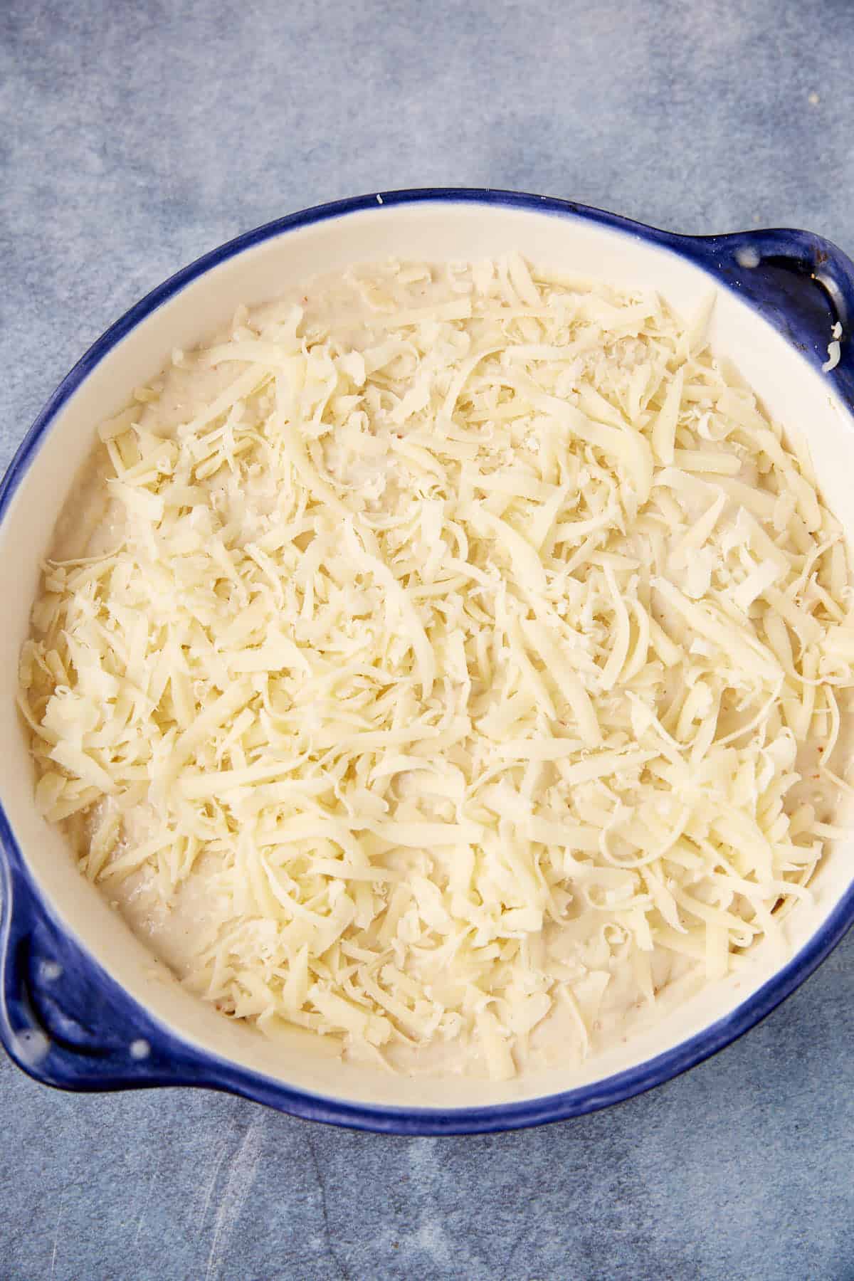 A round blue and white baking dish filled with a creamy chicken cordon bleu casserole, topped with a layer of shredded cheese, sits on a textured gray surface.