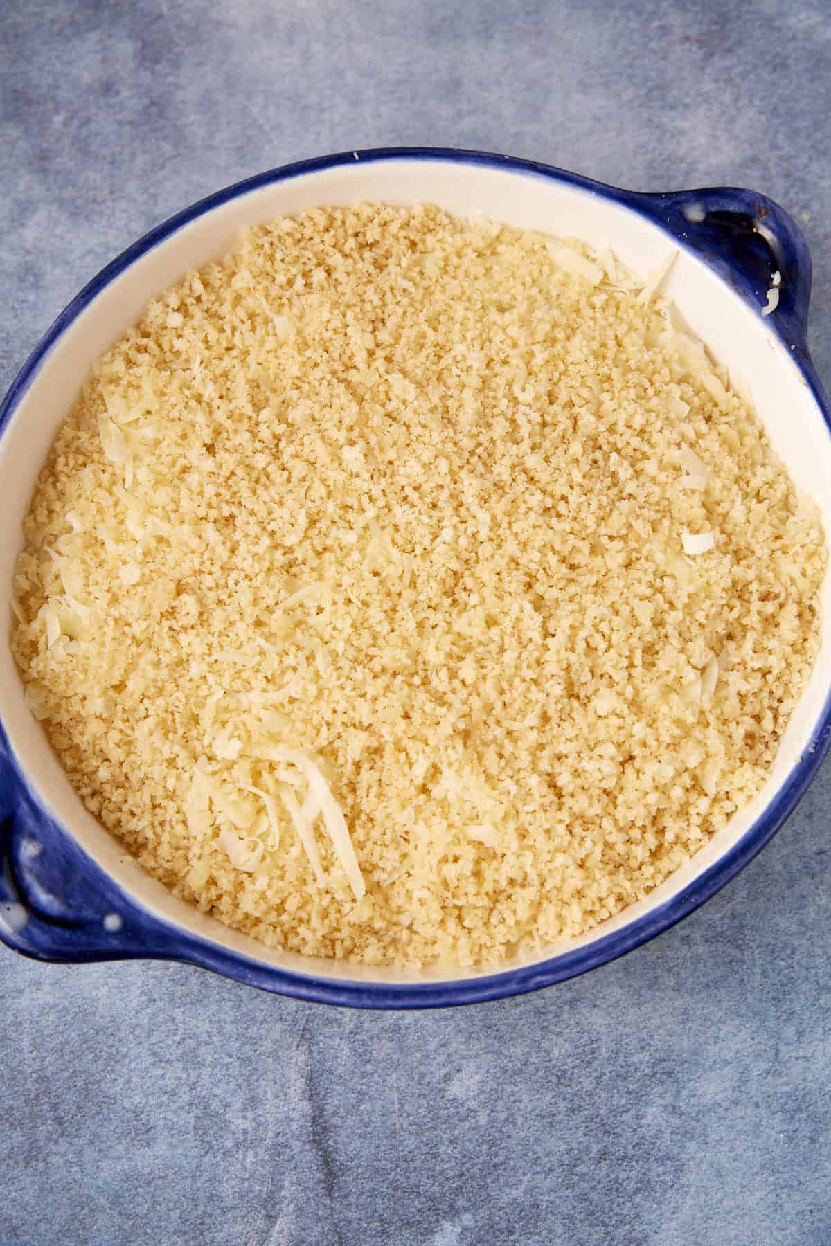 A round blue baking dish filled with a layer of shredded cheese and breadcrumbs, ready to be baked as a delicious chicken cordon bleu casserole, sits on a light blue textured surface.