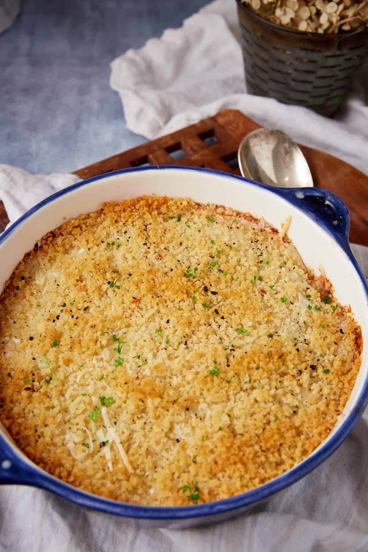 A baked chicken cordon bleu casserole with a golden, crispy breadcrumb topping in a blue-rimmed dish, garnished with herbs, sits on a white cloth with a spoon and a bowl of oats in the background.