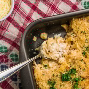 A baking dish of creamy, baked macaroni and cheese with a golden breadcrumb topping sits partially served with a spoon—evoking comfort food like chicken noodle casserole. Fresh parsley is sprinkled on top beside a bowl of shredded cheese on a red plaid tablecloth.