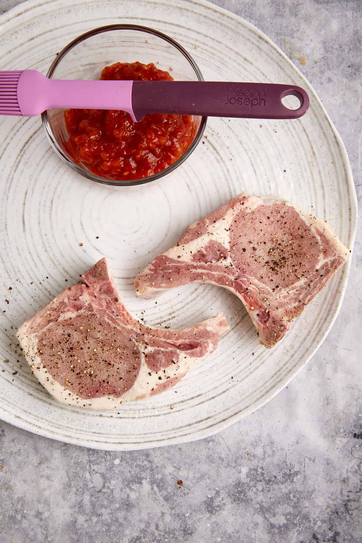 Two pork chops with salt and pepper next to a bowl of bbq sauce.
