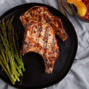 Two BBQ pork chops with peach sauce on a plate with asparagus.