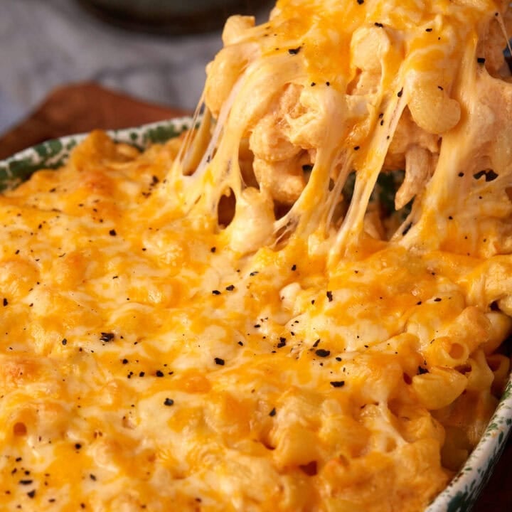 Close-up of buffalo mac and cheese with melted cheddar on top, being scooped out, showing gooey, stretchy cheese and golden, bubbly pasta with a sprinkling of black pepper.