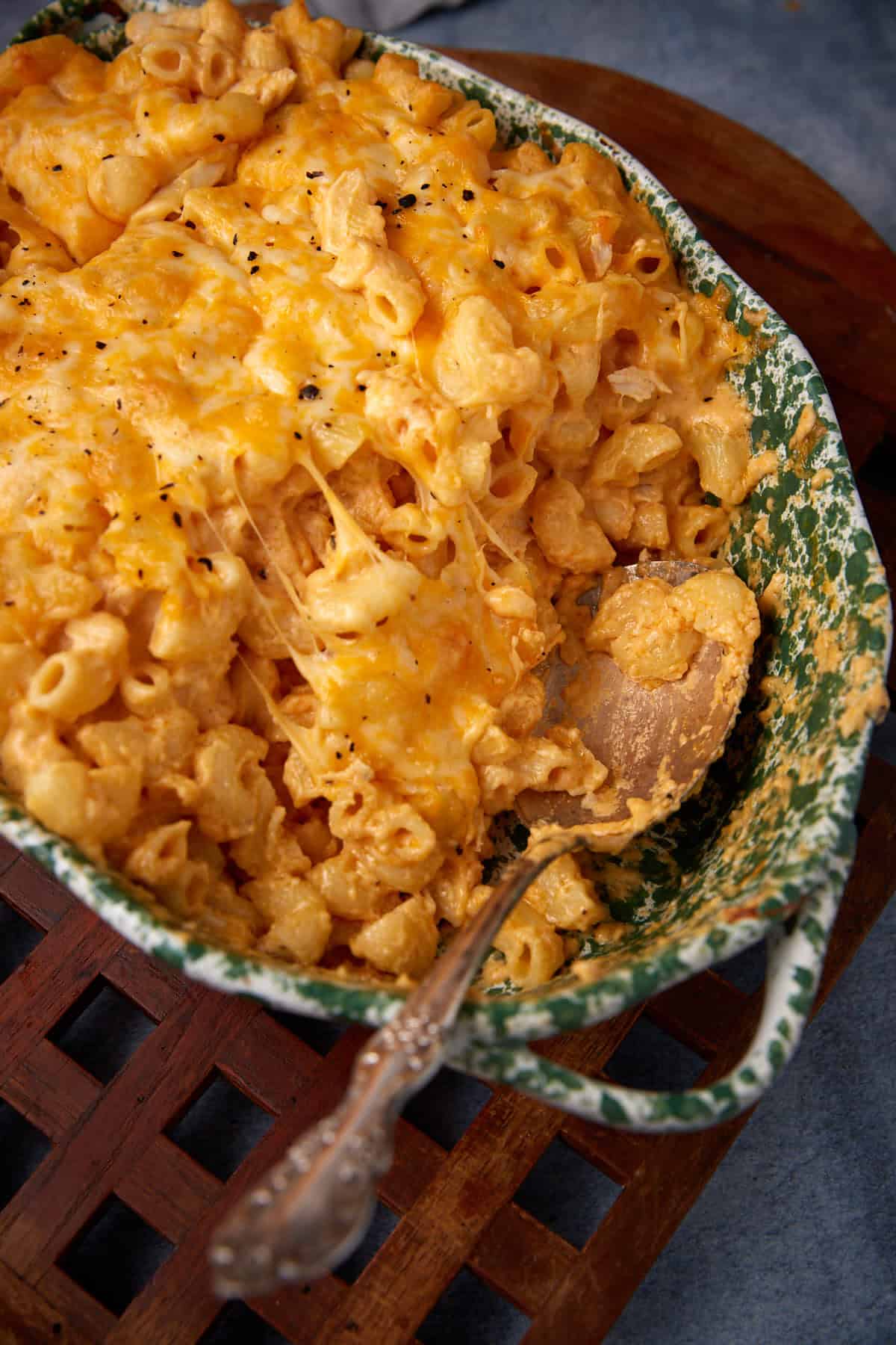 A green and white ceramic dish filled with buffalo mac and cheese, topped with melted cheese, with a serving spoon resting inside, placed on a wooden trivet.