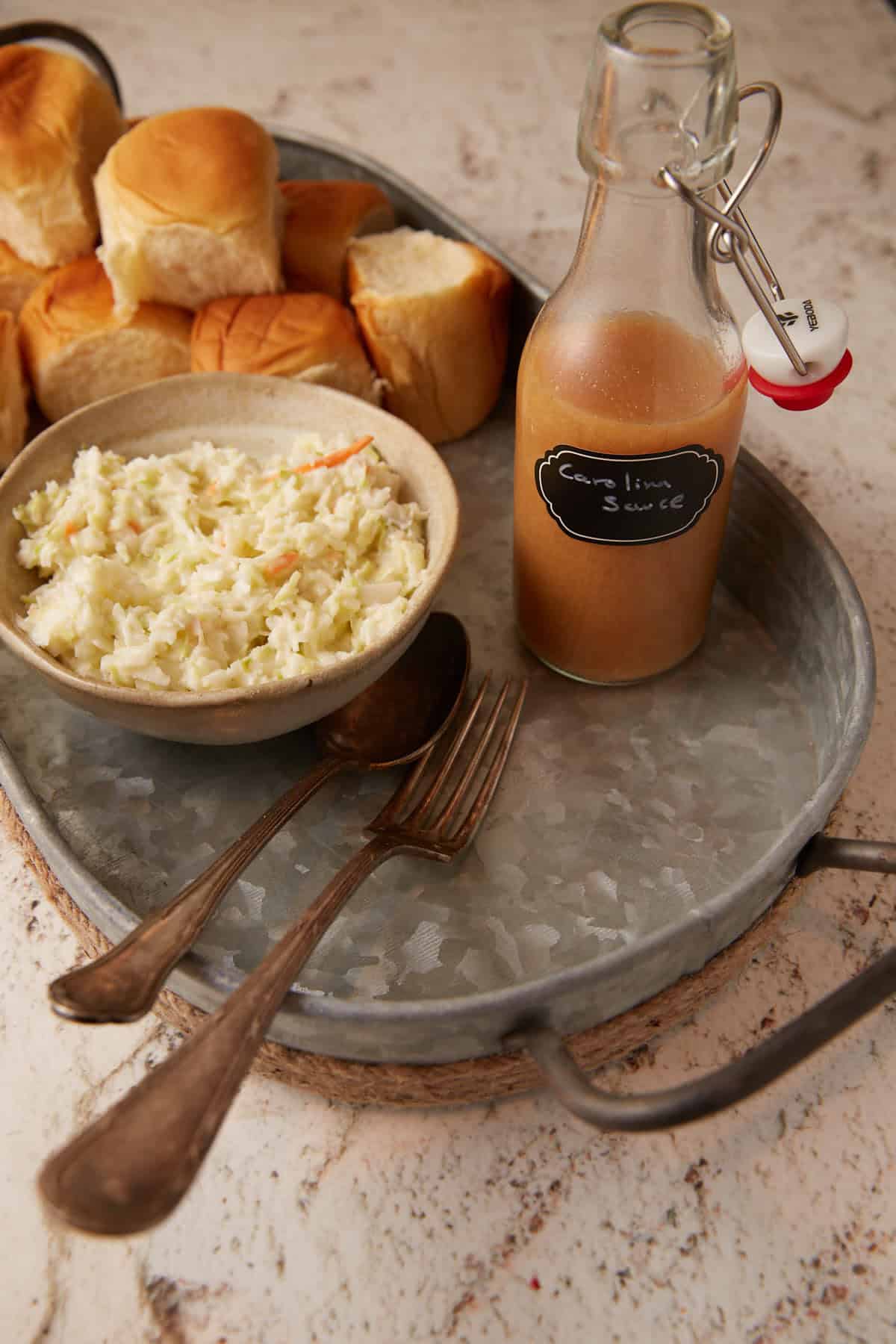 A tray with a bottle of carolina sauce next to rolls and cole slaw.