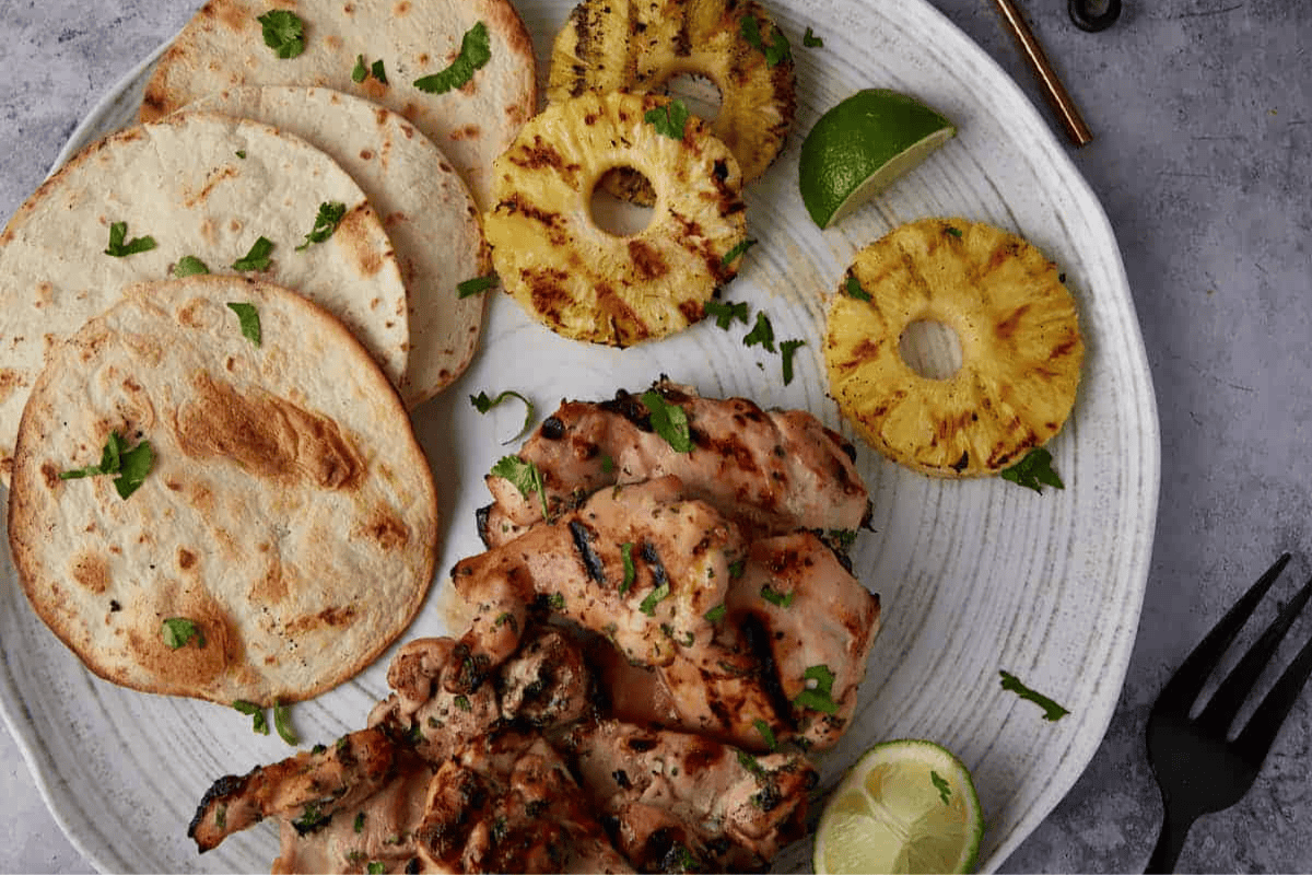 Grilled chicken on a platter with tortillas and pineapple slices.