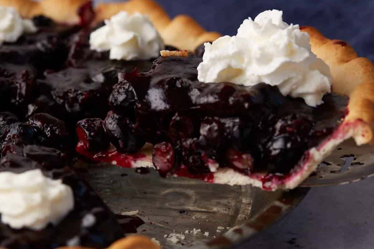 A freshly sliced blueberry pie topped with dollops of whipped cream is displayed on a metal pie dish. The deep purple filling contrasts with the golden crust, and the pie is partially cut, showcasing the juicy blueberries.