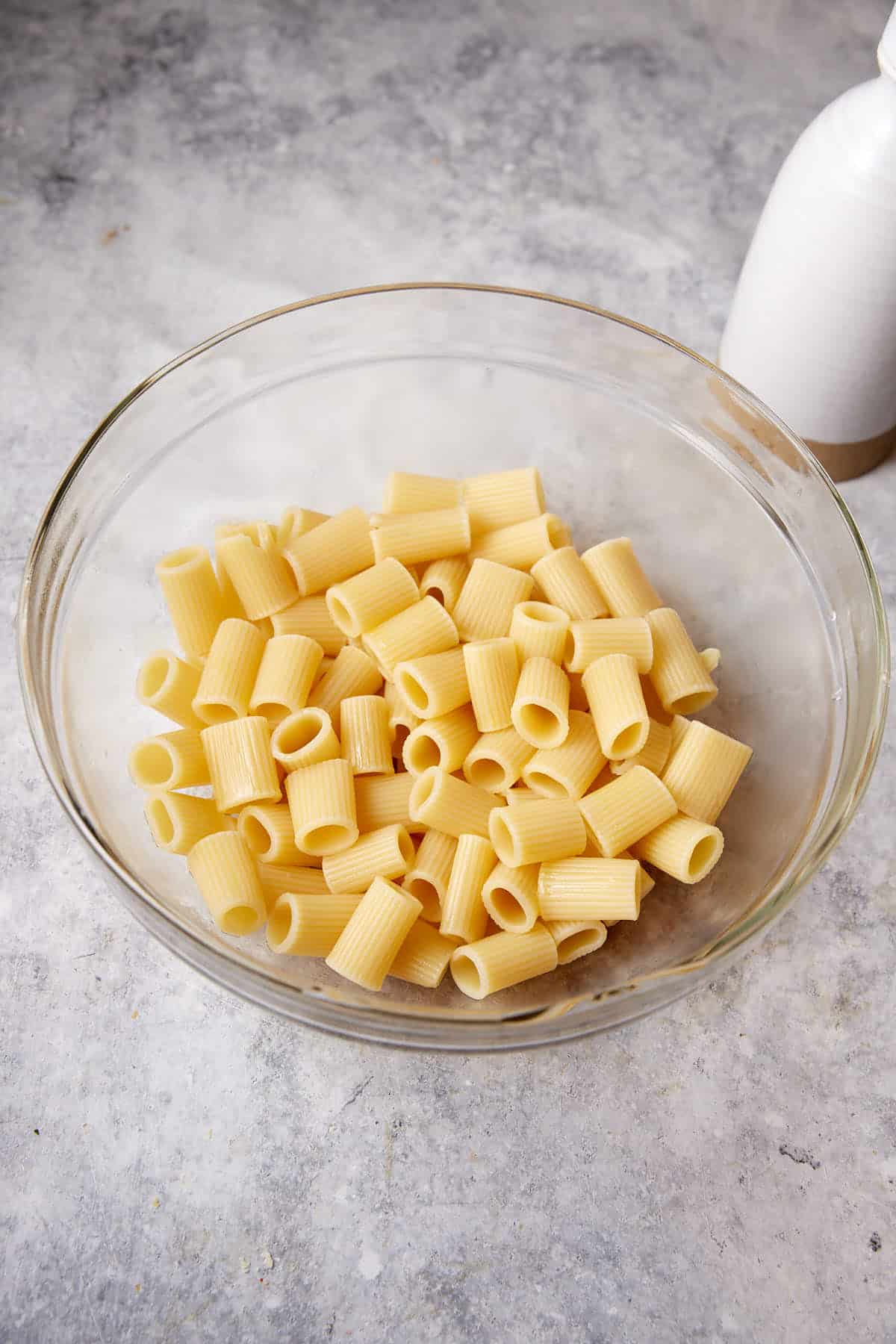 A glass bowl filled with uncooked rigatoni pasta sits on a gray, textured surface, ready for your favorite recipe like creamy chicken mac. Beside the bowl, the bottom portion of a white bottle is partially visible. The neutral background contrasts with the light yellow pasta.
