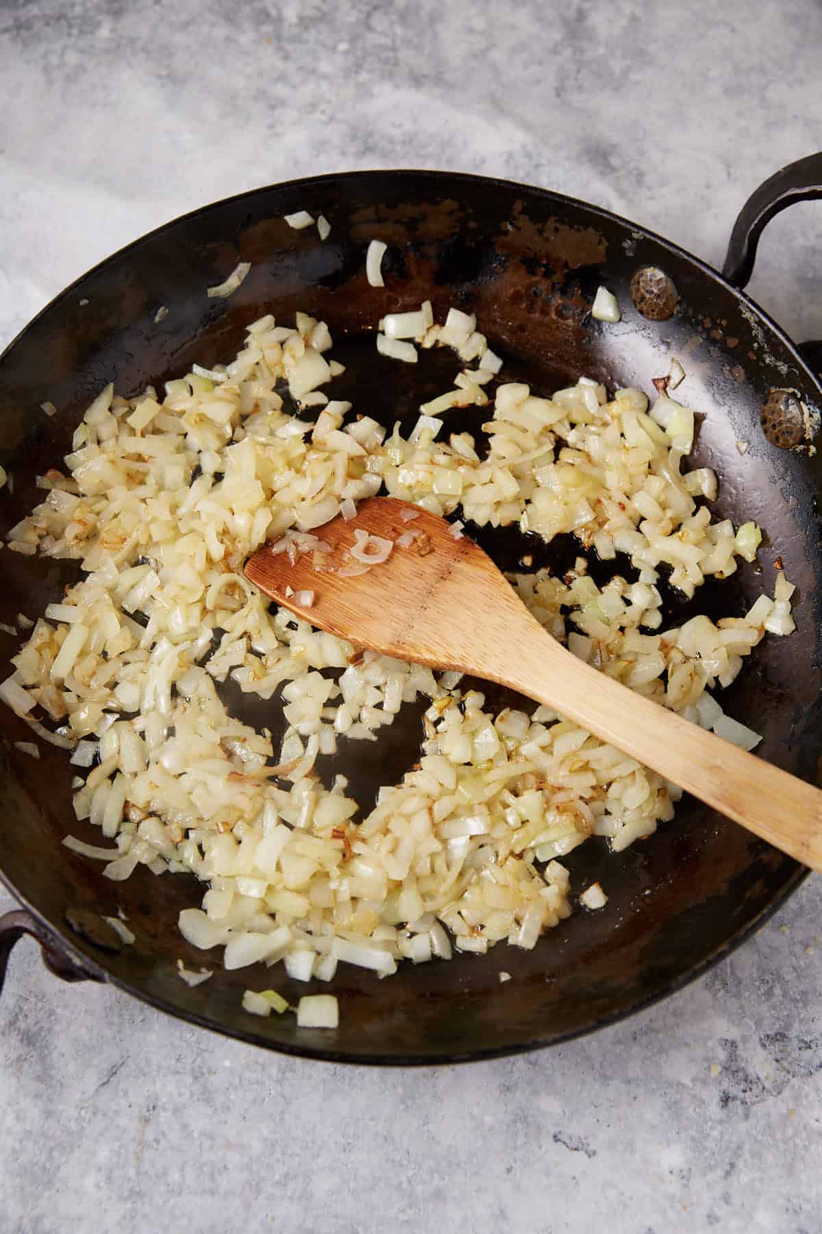A wooden spoon is placed in a black frying pan with chopped onions being sautéed. The onions are slightly browned and translucent, indicating they are being cooked. The frying pan, prepping the base for a delicious chicken mac, rests on a light-colored countertop.