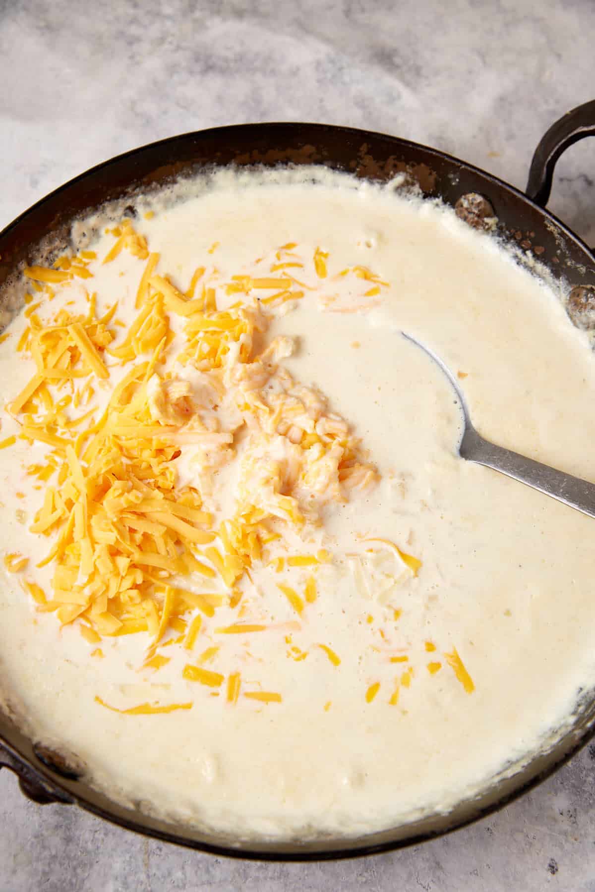 A frying pan filled with creamy white sauce and a pile of shredded cheese on top, perfect for chicken mac. A metal spoon rests in the pan, partially submerged in the sauce. The pan is set on a light, textured surface.
