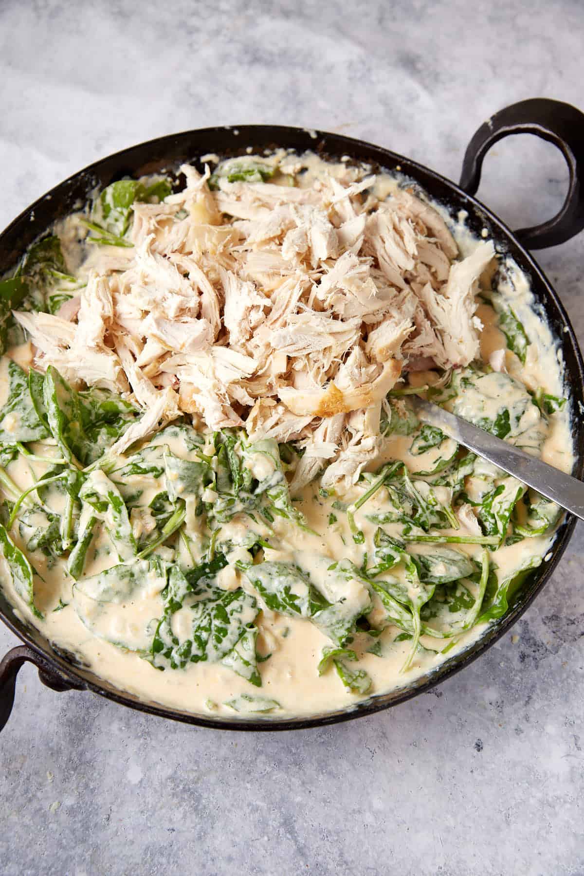 A black skillet filled with creamy white sauce mixed with shredded chicken and fresh spinach leaves, resembling a delectable chicken mac. A metal spoon rests in the skillet, showing a mixture of ingredients. The background is a grey textured surface.