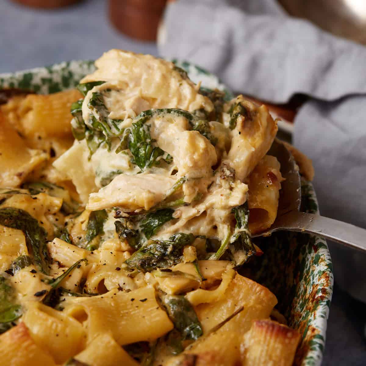 A spoon scooping out a portion of baked chicken Alfredo pasta from a casserole dish. The pasta is rich and creamy, mixed with spinach and chunks of chicken mac, and sprinkled with black pepper. The dish sits on a table, partially covered with a gray napkin.