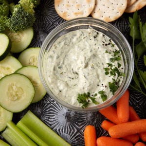 A bowl of creamy herb dip is surrounded by fresh vegetables, including broccoli, cucumber slices, baby carrots, celery sticks, and round crackers. The dip is garnished with chopped herbs and black pepper. The background features a textured, dark surface.