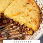 A close-up of a cornbread hamburger pie with a portion cut out, exposing the savory ground meat filling underneath. The pie, topped with golden cornbread, sits in a floral-patterned dish. A serving utensil is partially visible, indicating the hamburger pie is being served.