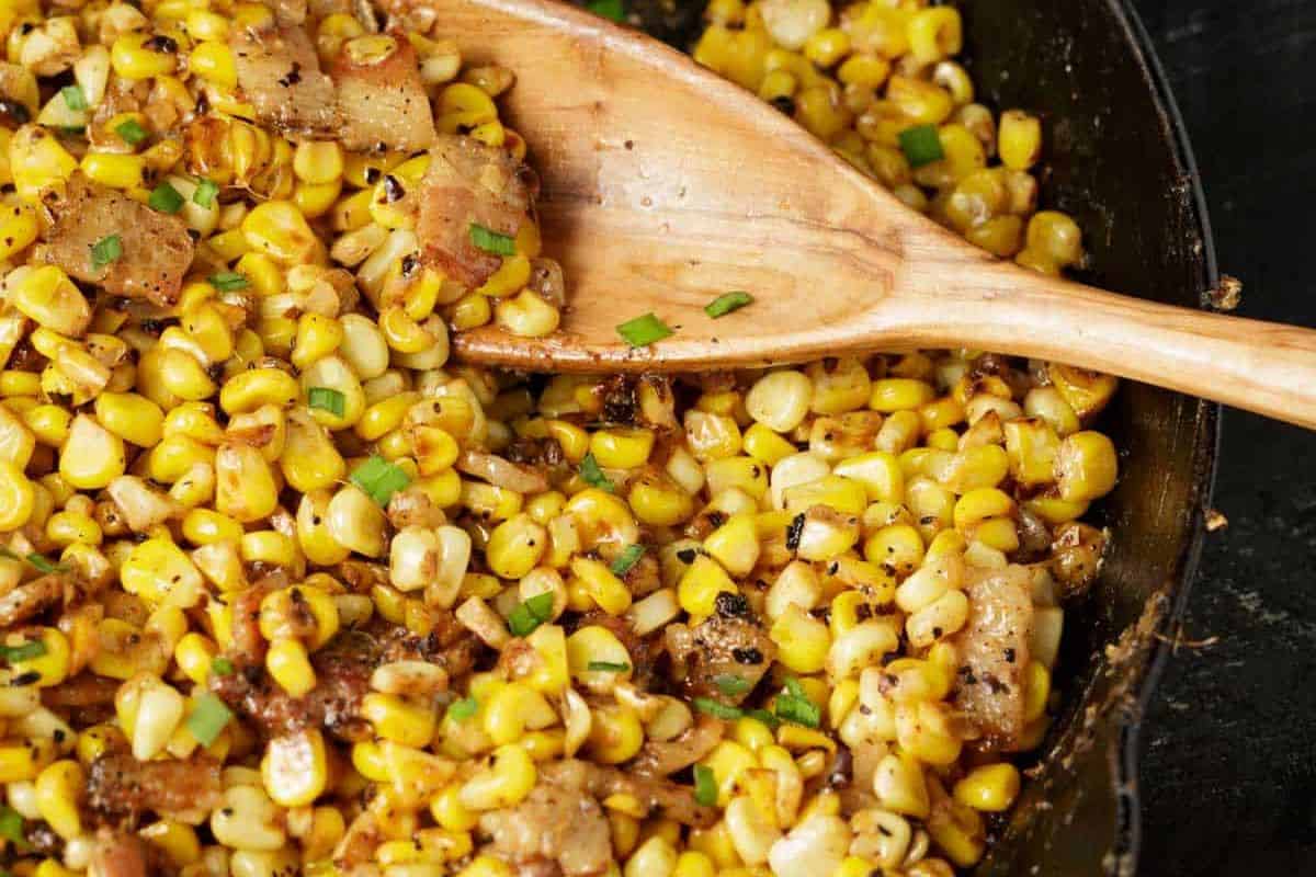 A close-up of a skillet filled with freshly cooked corn sautéed with bits of onions, green herbs, and seasonings. A wooden spoon is stirring the mixture, which appears golden and slightly charred in some spots, highlighting a delicious and inviting texture.