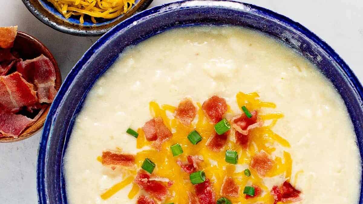 A bowl of creamy potato soup topped with shredded cheddar cheese, crispy bacon pieces, and chopped green onions. Next to the bowl are small dishes containing more shredded cheese and bacon bits. All items are on a white surface.