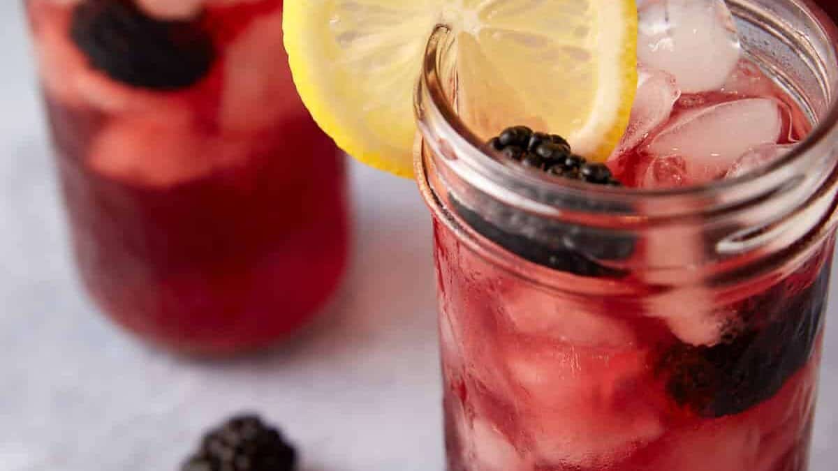 A close-up of a refreshing berry drink in a mason jar filled with ice cubes. The drink is a deep red color, and it is garnished with a slice of lemon and a blackberry. Another similar drink is visible in the background, along with a scattered blackberry.