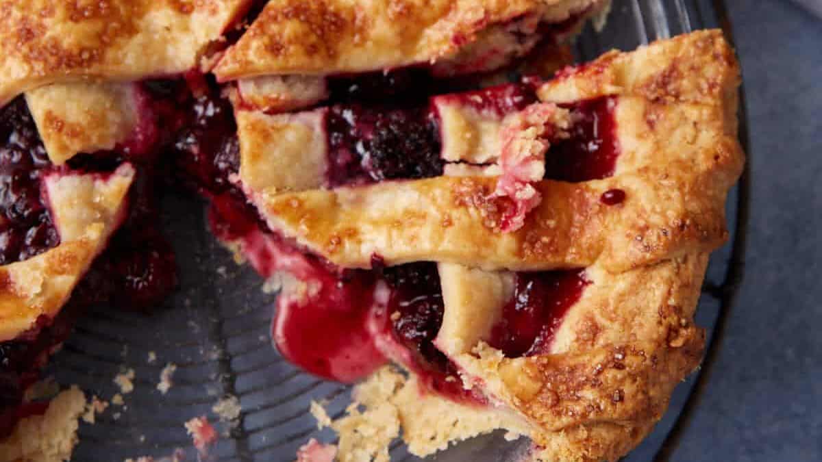 A top-down view of a partially eaten blackberry pie with a lattice crust on a wire cooling rack. The pie has a crispy golden-brown crust with juicy, dark purple filling. A bowl of whipped cream is placed nearby. Crumbs and a piece of pie are visible on the table.