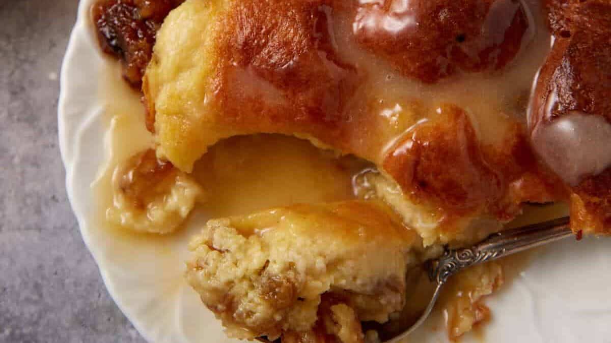 A close-up of a dessert on a white plate. The dessert appears to be bread pudding, covered in a rich, glossy sauce. A spoon with a bite of the pudding rests on the plate. In the background, there are additional pieces of dessert and vintage cutlery on a gray surface.
