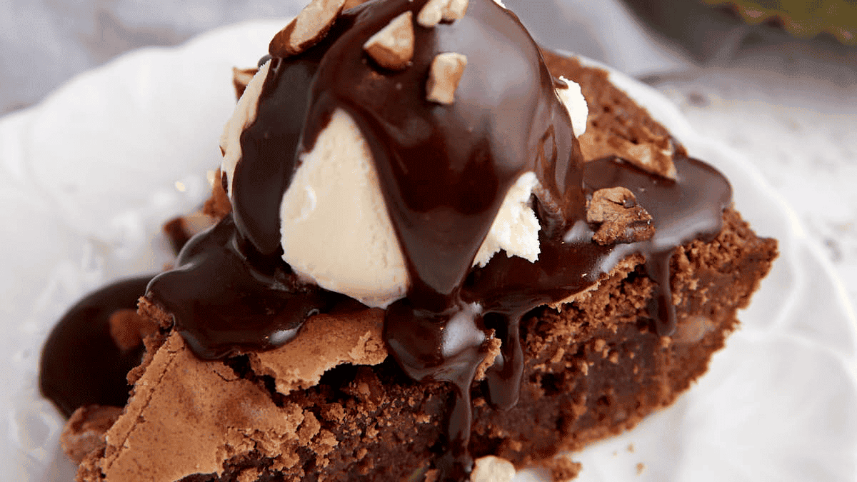 A slice of chocolate pie topped with a scoop of vanilla ice cream and drizzled with chocolate sauce. Garnished with chopped nuts, this southern dessert sits on a white plate, with a grey fabric napkin slightly visible in the background.