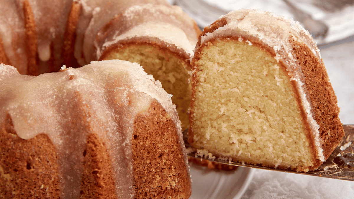 A close-up of a sliced bundt cake with a light glaze. The southern dessert is golden brown with a moist, dense interior. One piece is cut and raised on a cake server, revealing the soft cake texture inside. The background includes a white surface and blurred utensils.