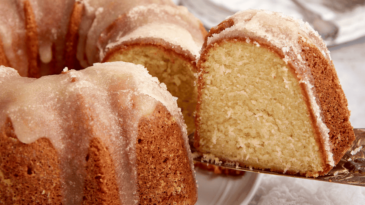 A close-up of a sliced bundt cake with a light glaze. The southern dessert is golden brown with a moist, dense interior. One piece is cut and raised on a cake server, revealing the soft cake texture inside. The background includes a white surface and blurred utensils.
